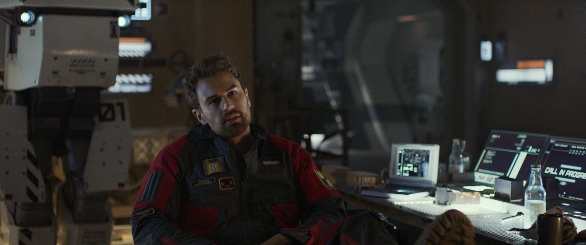 Theo James leans back with his feet on his messy tech-desk and his prototype robot J1 in the background in Archive.